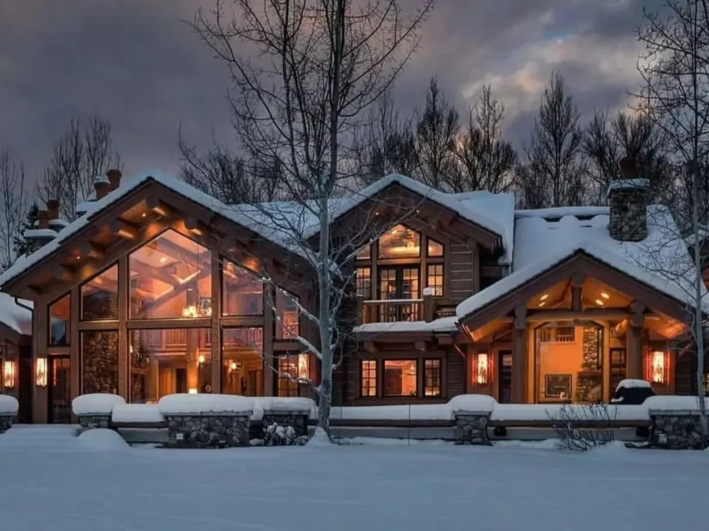 Embracing Grandeur: 25 Inspiring Ideas for Large Log Cabin Homes 13 A large log cabin home covered in snow at dusk, with warm light shining through its large windows.