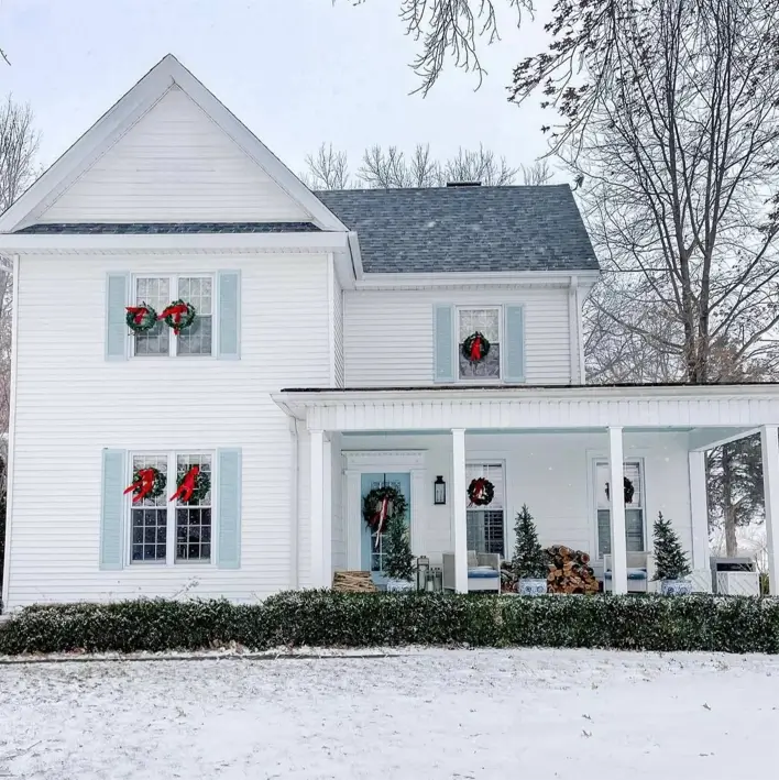 A classic off-white farmhouse in the snow, decorated for Christmas with wreaths on its light blue shutters.