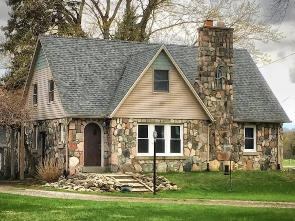 A rustic cottage with rounded river stone walls and a large stone chimney featuring a monogram.