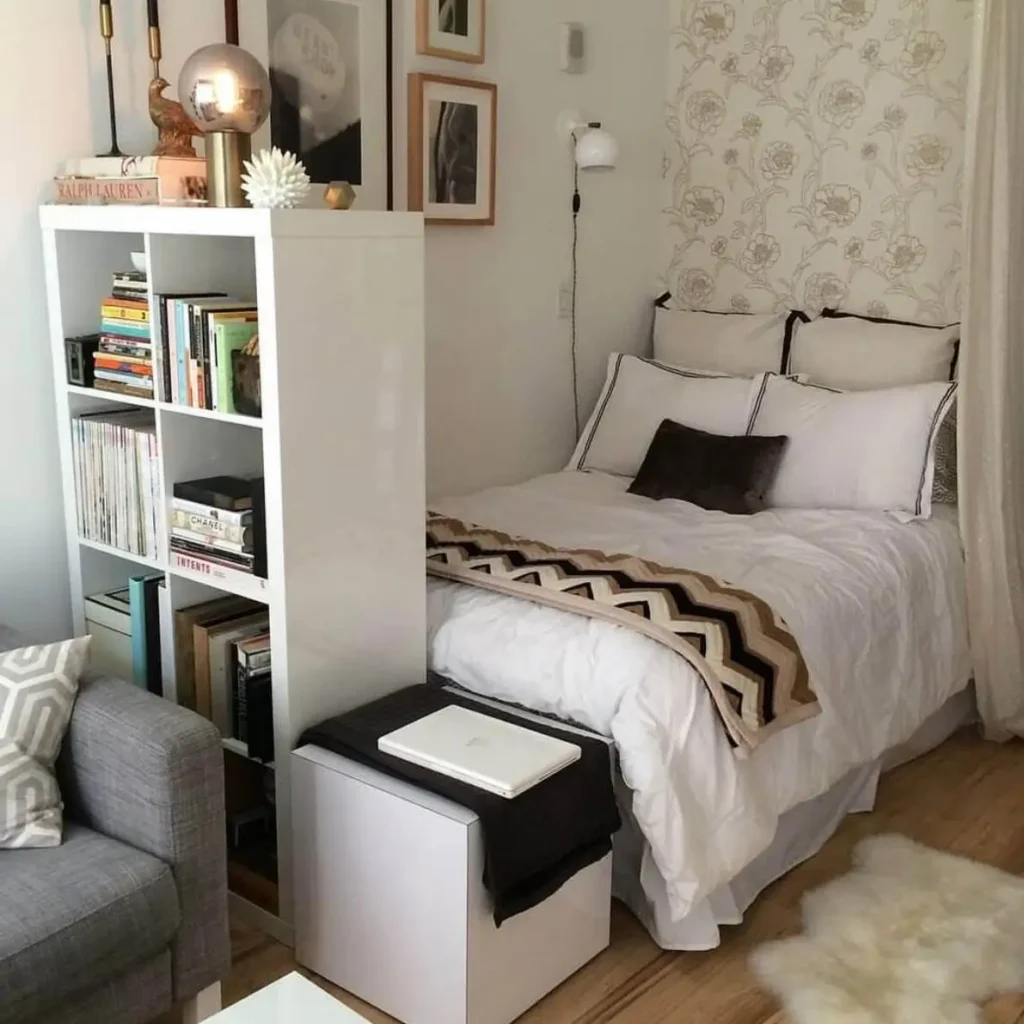 A small bedroom interior design featuring a white bookshelf used as a room divider next to a bed with a patterned throw blanket.