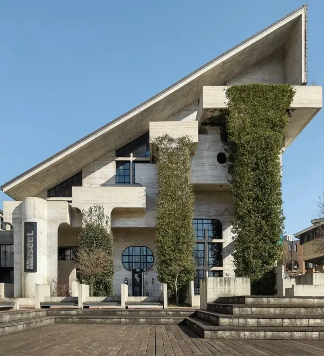 A monumental brutalist building with a dramatic, sharply sloped concrete roof and tall structural columns covered in lush green ivy.