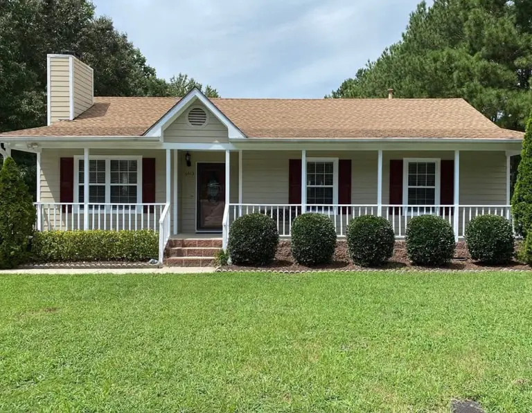 A welcoming beige ranch house with a full-length white porch and deep red shutters.