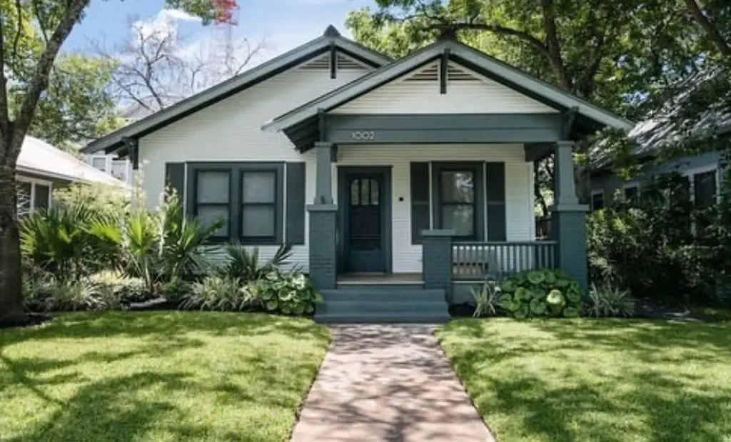 A white craftsman bungalow with a dark teal-gray front porch, matching shutters, and a well-manicured lawn.