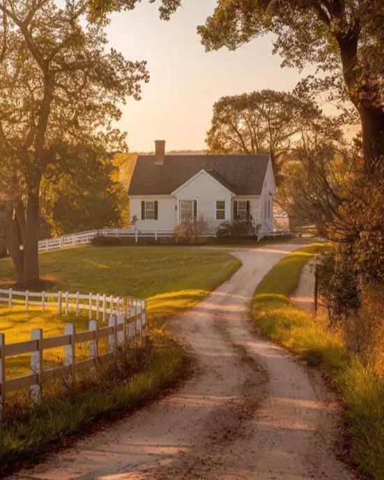 An idyllic country house design featuring a simple white farmhouse at the end of a winding dirt road at sunset.