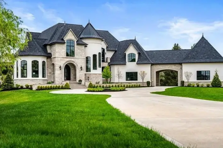 20 Stone Mansion Ideas for Classic & Modern Designs 13 A modern stone mansion with white walls, a dark roof, a turret, and a covered driveway, set on a manicured green lawn.