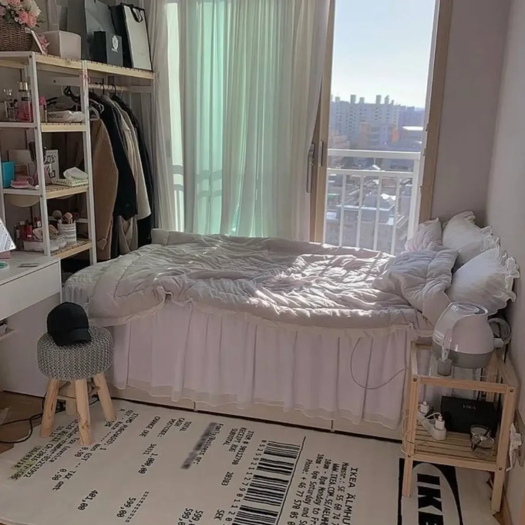 A small, bright bedroom with open-concept shelving for clothes, a bed with a white ruffled skirt, and a unique receipt-themed area rug.