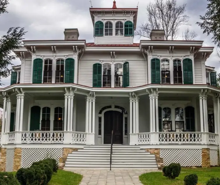 A grand historic country house design with white siding, vibrant green shutters, a wraparound porch, and a rooftop cupola.