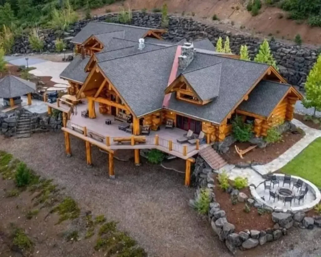 Embracing Grandeur: 25 Inspiring Ideas for Large Log Cabin Homes 15 An aerial view of a large log cabin featuring an elevated wraparound deck on log stilts and a separate sunken stone fire pit.