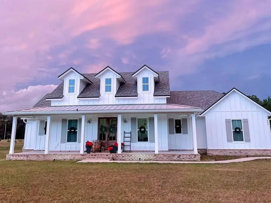 An off-white farmhouse exterior with three dormer windows and a brick porch base, set against a dramatic pink sunset sky.