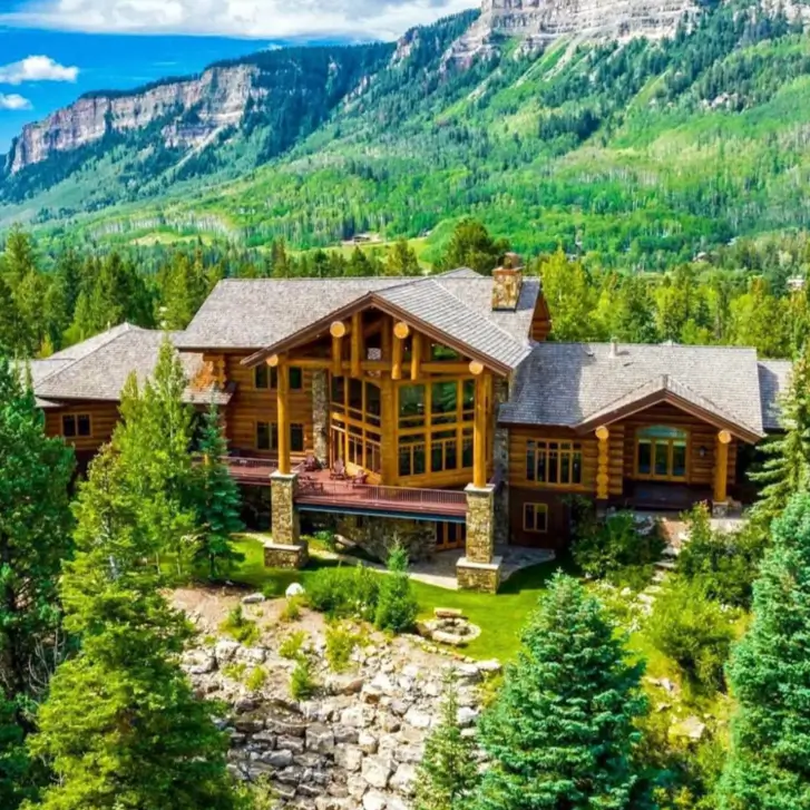 Embracing Grandeur: 25 Inspiring Ideas for Large Log Cabin Homes 16 A large log cabin home nestled in a forest at the base of a dramatic mountain range under a blue sky.