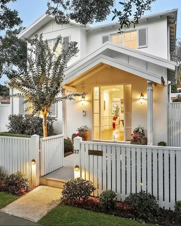 A charming off-white farmhouse with a well-lit gabled entryway and a white picket fence at dusk.