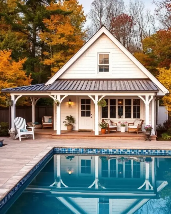 A white farmhouse-style pool house with a curved metal roof, reflected in a clear blue pool and surrounded by colorful autumn trees.