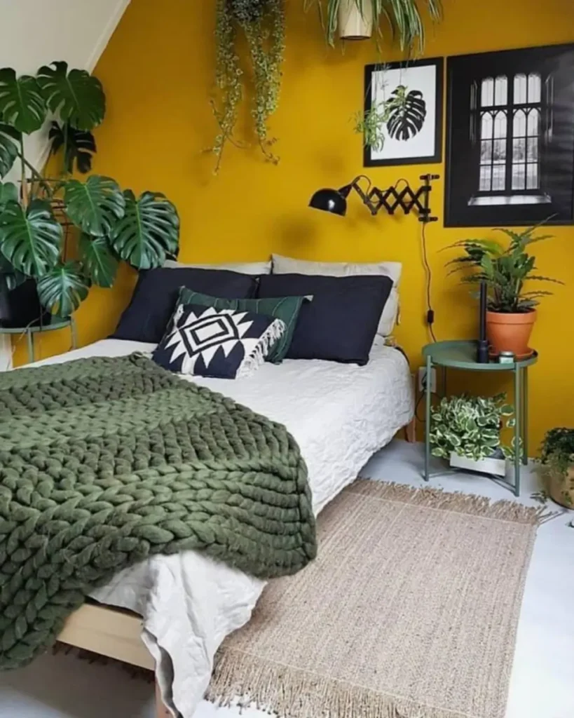 A bedroom with a bold mustard yellow wall, a bed featuring a large olive green chunky knit throw, and many plants.