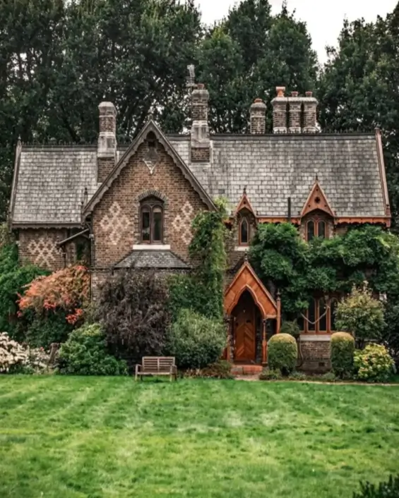 A Gothic Revival country house design featuring detailed brickwork, pointed arches, and a slate roof, surrounded by lush greenery.