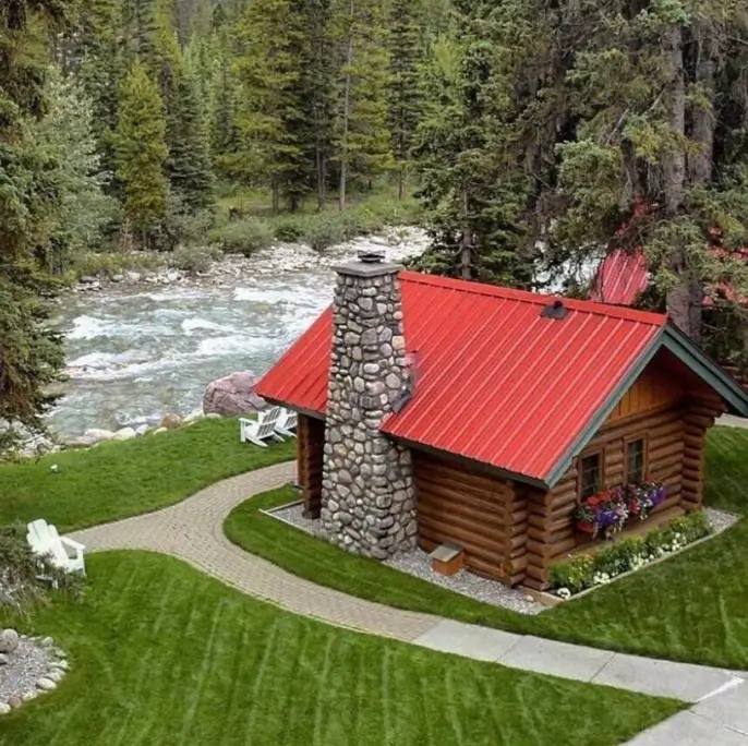 Small Log Cabins: 36 Ideas for Cozy Living & Enduring Charm 17 A small log cabin featuring a bright red metal roof and a large stone chimney, set on a green lawn next to a flowing river.