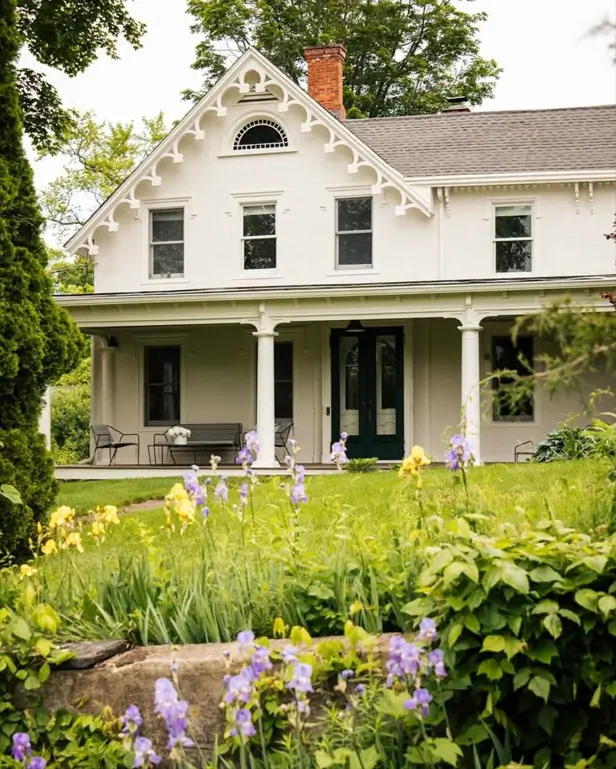 An off-white Victorian farmhouse with decorative gable trim and a lush flower garden in the foreground.