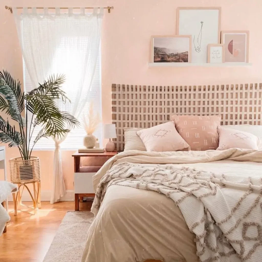 A sunny boho bedroom with light pink walls, a woven headboard, and a white shelf displaying framed art prints.