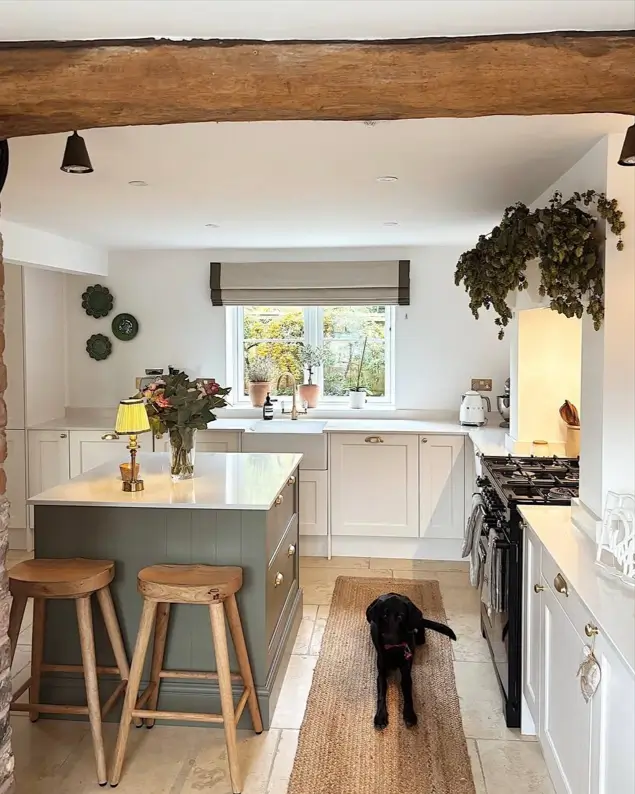 A modern rustic kitchen with a sage green island, white cabinetry, a jute runner, and a prominent overhead wooden beam.