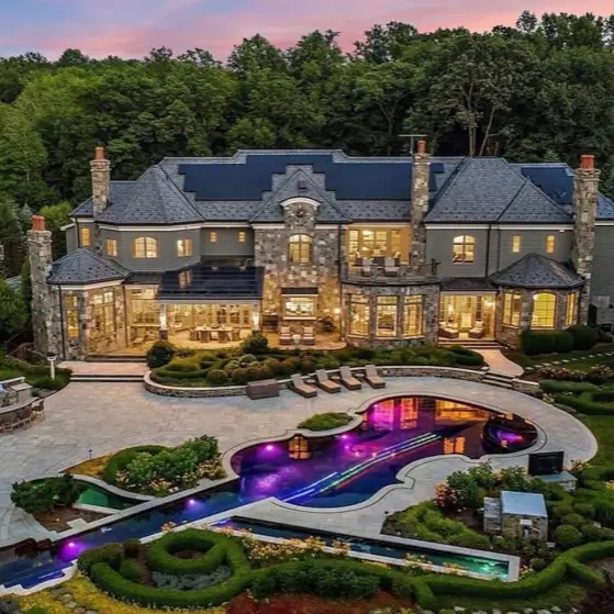 20 Stone Mansion Ideas for Classic & Modern Designs 17 The rear exterior of a modern stone mansion at twilight, featuring an expansive patio and a uniquely shaped, illuminated swimming pool.