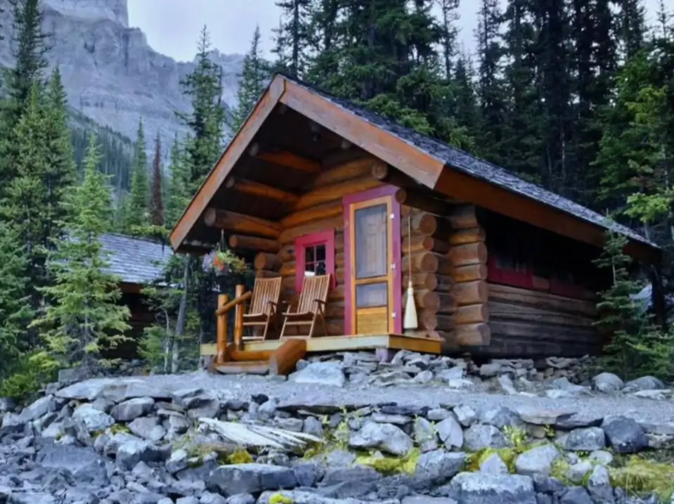 60 Charming Small Log Cabin Ideas to Inspire Your Rustic Retreat 17 A rustic small log cabin home situated on a rocky shoreline with pine trees and mountains behind it.