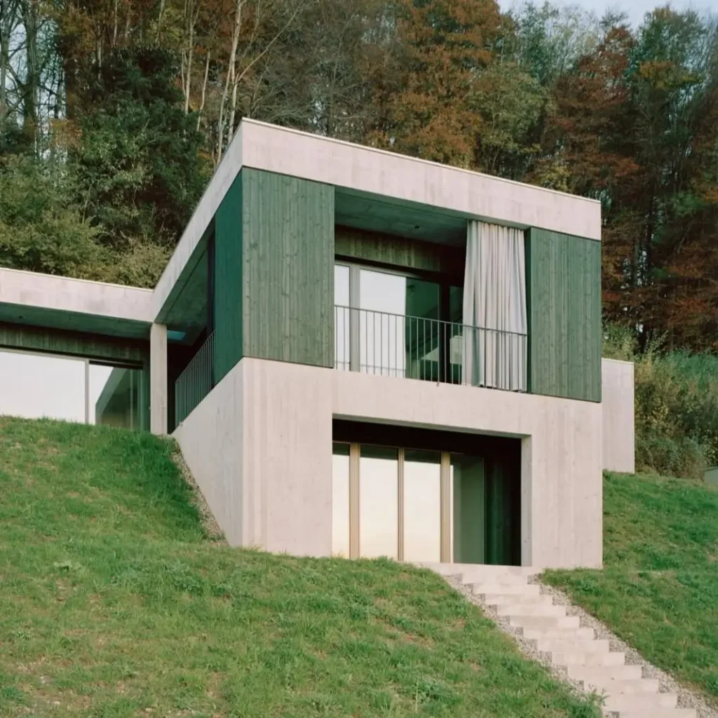 A contemporary concrete home terraced into a green hill, featuring dark wood panels on the upper level.