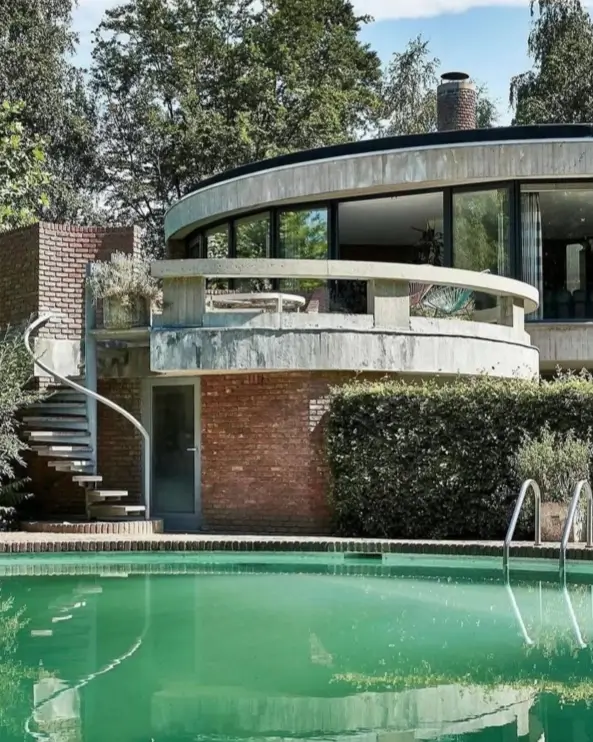 A classic brutalist home design combining a curved concrete balcony with the warm, earthy texture of red brick walls by a poolside.