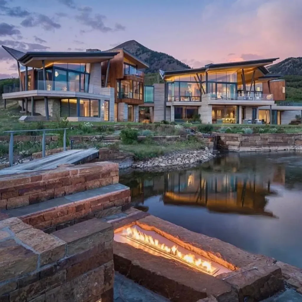 16 Mountain Mansions That Define Luxury Alpine Living 20 Dramatic outdoor living space of a modern mountain mansion featuring a stone fire pit integrated at the edge of a tranquil pond at dusk.