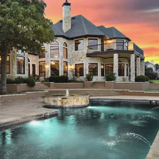 20 Stone Mansion Ideas for Classic & Modern Designs 18 The backyard of a modern stone mansion at sunset, with an illuminated pool and spa in the foreground and a large covered balcony.