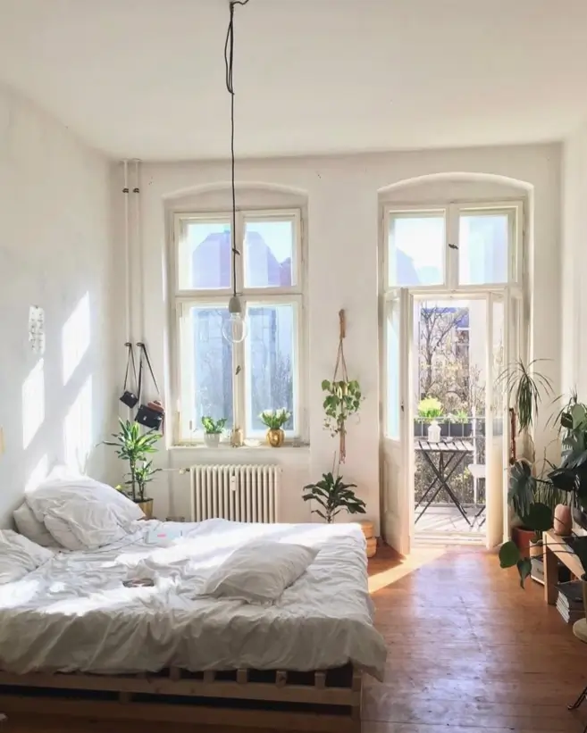 A bright, airy small bedroom with a low pallet bed, white linens, hardwood floors, and abundant sunlight from tall windows.