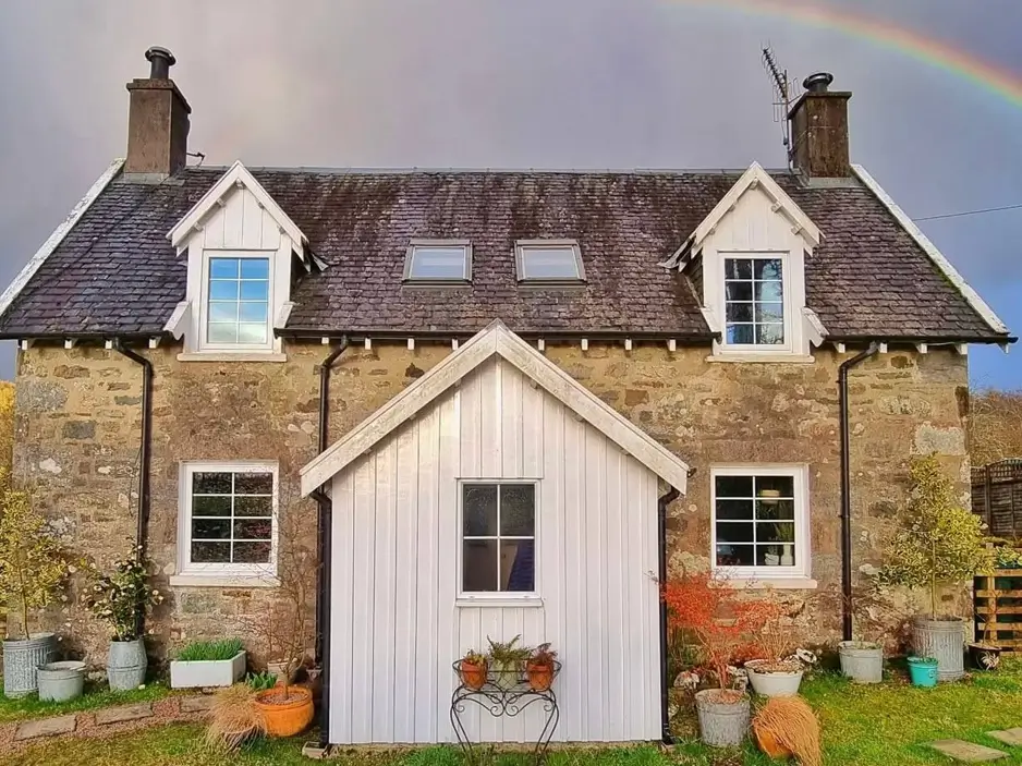 A symmetrical stone cottage with a white wooden vestibule and a rainbow in the sky.