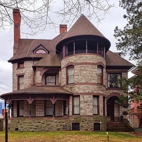 20 Stone Mansion Ideas for Classic & Modern Designs 19 A historic stone mansion featuring a rounded turret with an upper balcony and a wraparound porch with decorative trim.