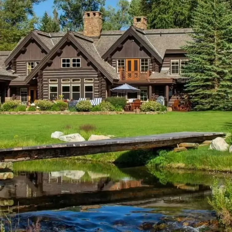 Embracing Grandeur: 25 Inspiring Ideas for Large Log Cabin Homes 20 A dark-stained log cabin with a large lawn, viewed from across a gentle stream with a simple wooden footbridge.