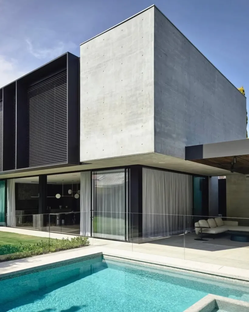 A modern brutalist house featuring a massive, cantilevered concrete volume, contrasted with sleek black window frames and a tranquil swimming pool.