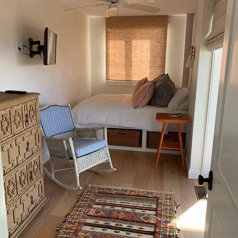 A small cozy bedroom with a bed tucked into a nook, featuring under-bed storage with wicker baskets and a carved wooden dresser.