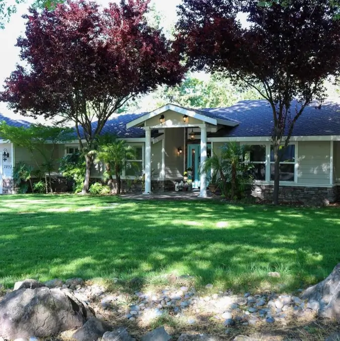 A taupe ranch house with stone accents, a bright teal front door, and prominent dark red trees.