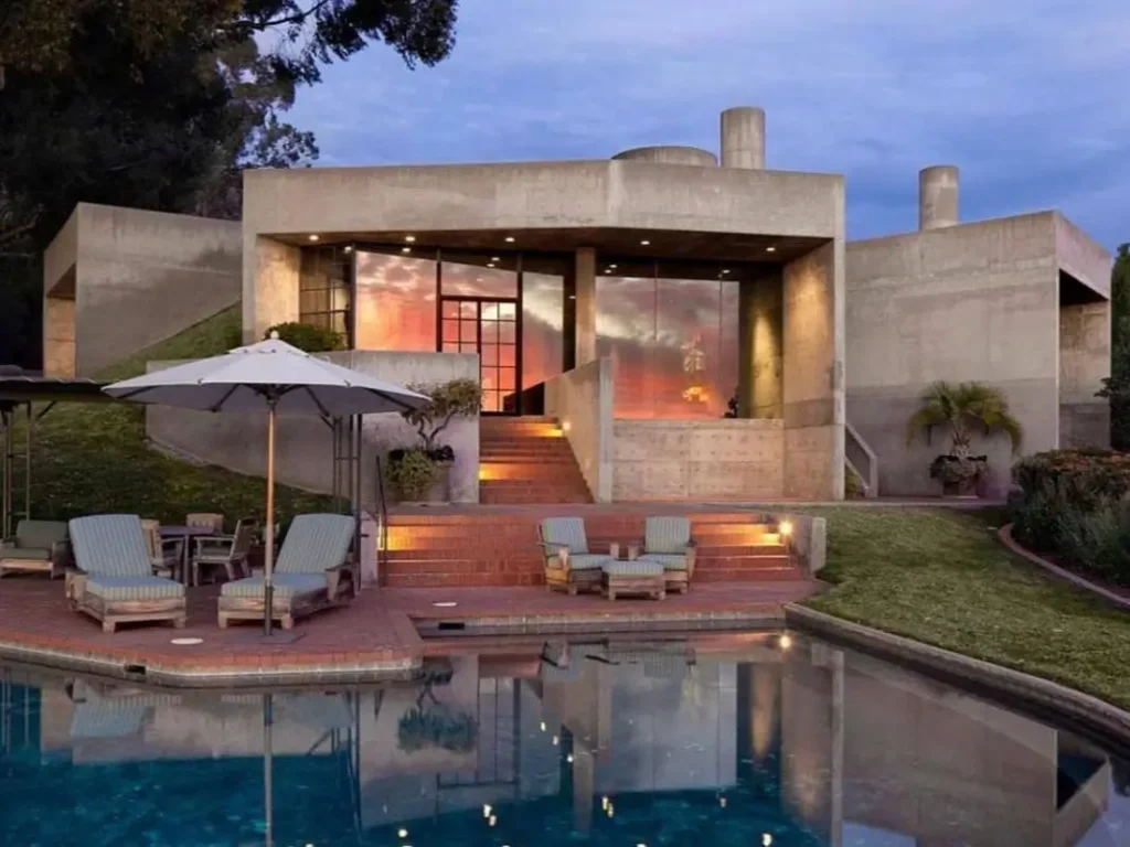 A geometric concrete brutalist house at dusk, its warm interior lights and the sunset reflecting beautifully in the large glass windows overlooking a pool.