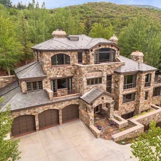20 Stone Mansion Ideas for Classic & Modern Designs 20 A multi-level modern stone mansion nestled into a lush, green hillside, featuring arched wooden garage doors and large windows.
