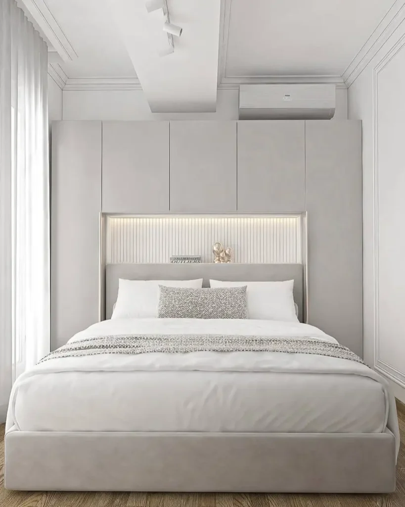A small, modern bedroom with a custom light gray built-in headboard and storage unit featuring soft backlighting.