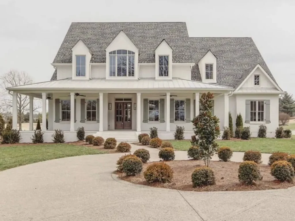 A stately off-white brick farmhouse with a circular driveway, manicured shrubs, and soft green shutters.