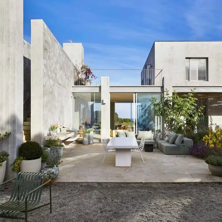 A sun-drenched patio of a concrete brutalist house, designed as a seamless outdoor living room with a dining set and comfortable sofa.