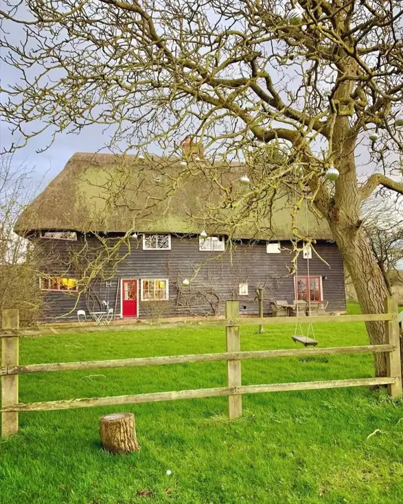 Dark wood British country house with a thatched roof and a bright red door, seen from behind a wooden fence.