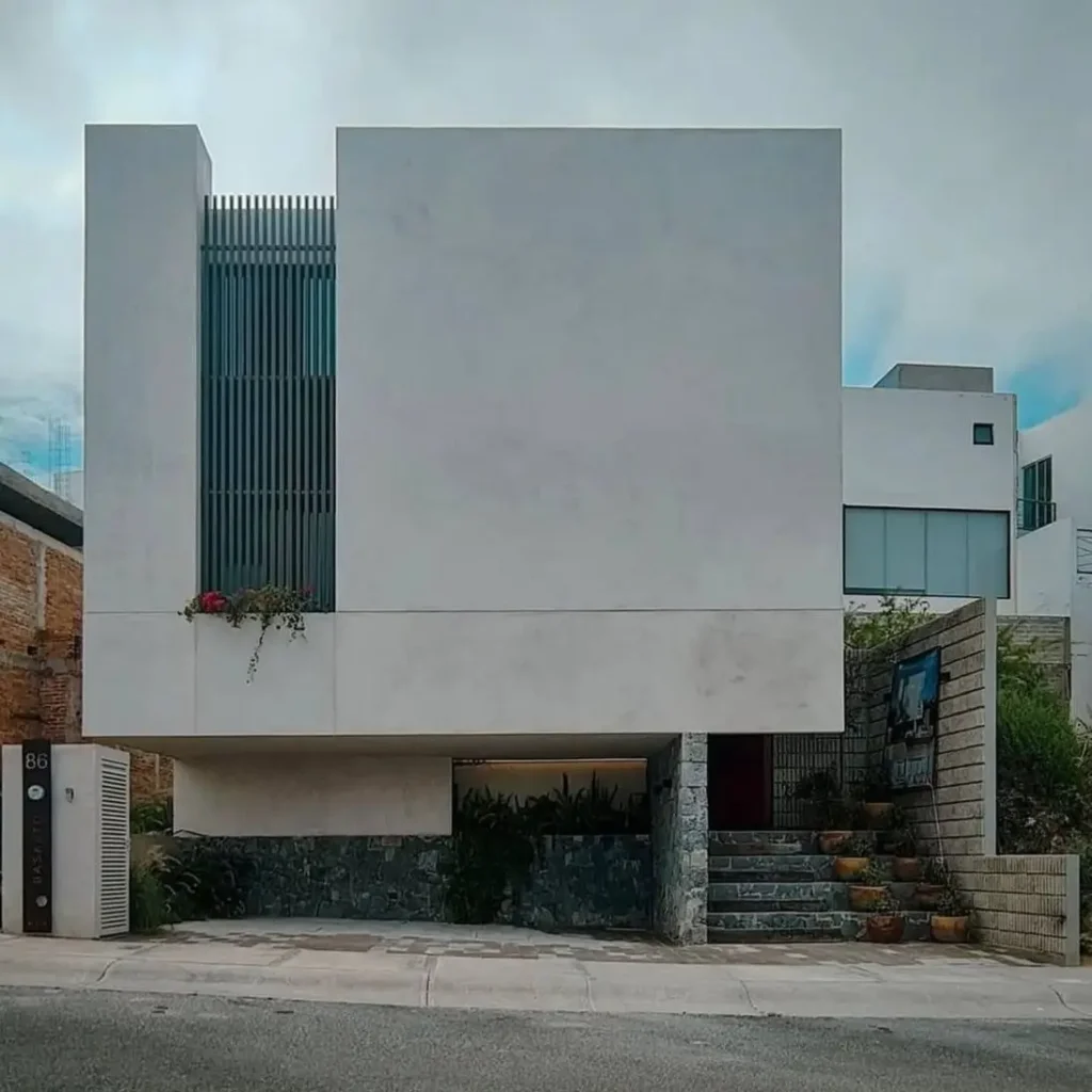 A modern white concrete house designed as a floating cube over a dark stone-clad ground floor.