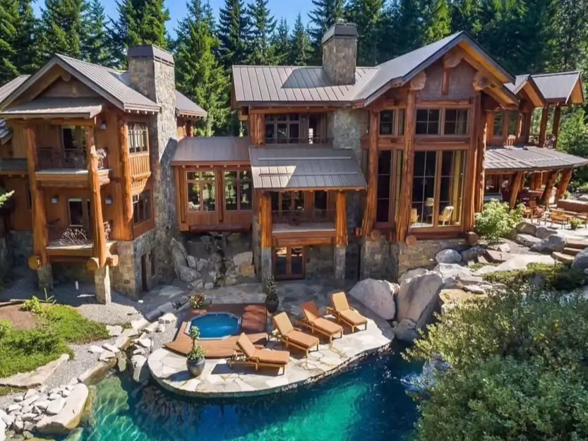 Embracing Grandeur: 25 Inspiring Ideas for Large Log Cabin Homes 22 A modern log cabin home with a stone and wood exterior, featuring a lagoon-style pool and hot tub built into a rocky hillside.
