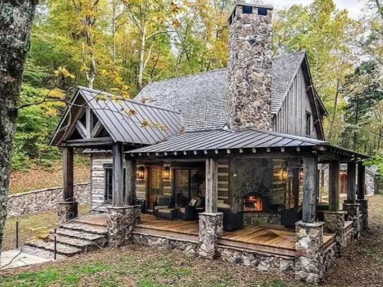 Small Log Cabins: 36 Ideas for Cozy Living & Enduring Charm 22 A small rustic log cabin with a large stone chimney and a covered porch featuring an outdoor fireplace.