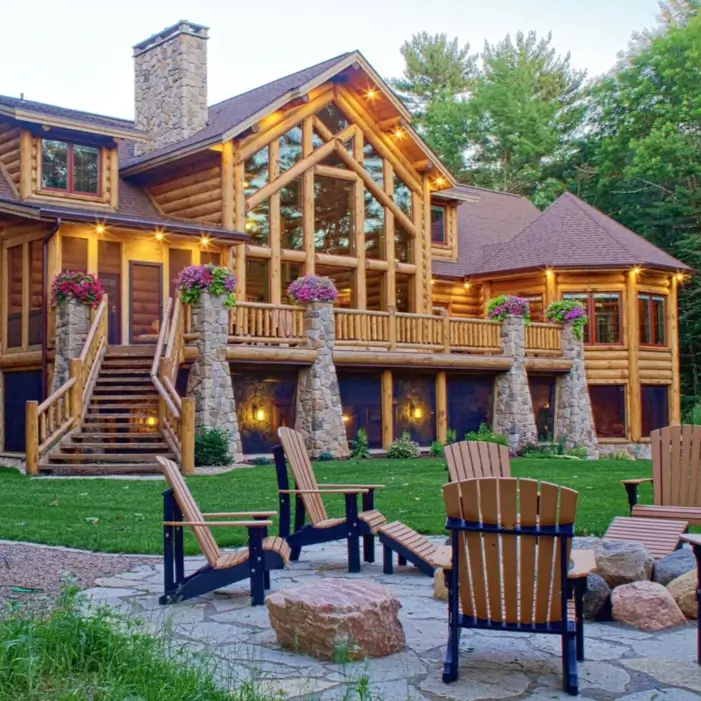 Embracing Grandeur: 25 Inspiring Ideas for Large Log Cabin Homes 5 A big log cabin home featuring a prominent stone foundation, a second-story deck, and a stone fire pit with chairs.