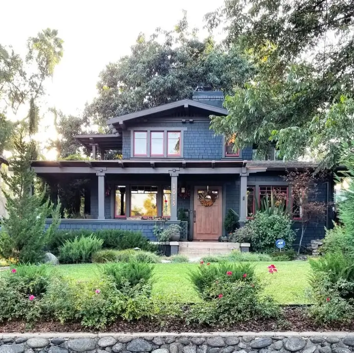 A dark blue craftsman house exterior with contrasting reddish-brown window trim and a wide front porch surrounded by mature landscaping.