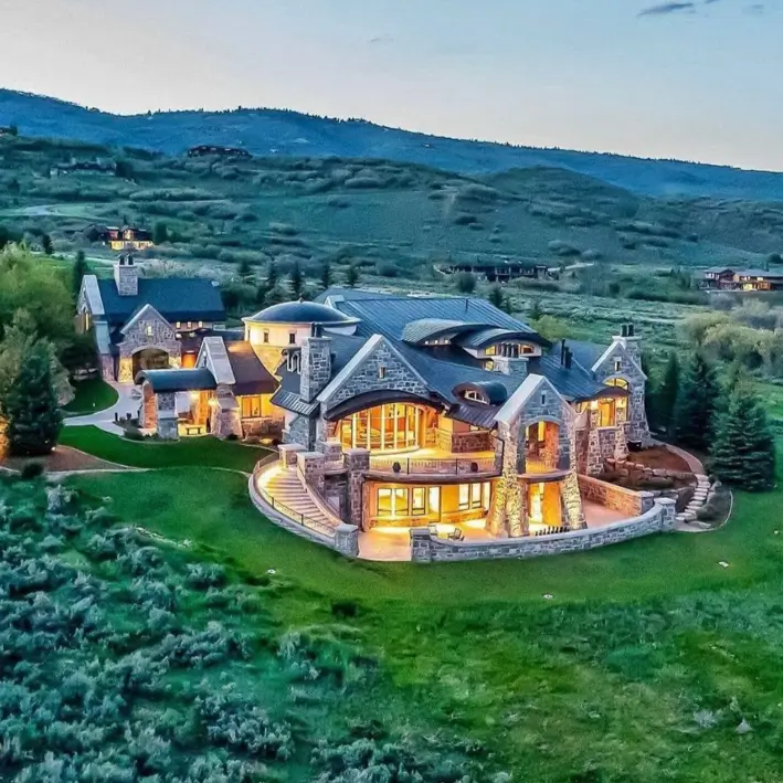 16 Mountain Mansions That Define Luxury Alpine Living 6 Sprawling luxury mountain mansion made of grand stonework, with warm interior lights glowing at dusk on a rolling green hill.