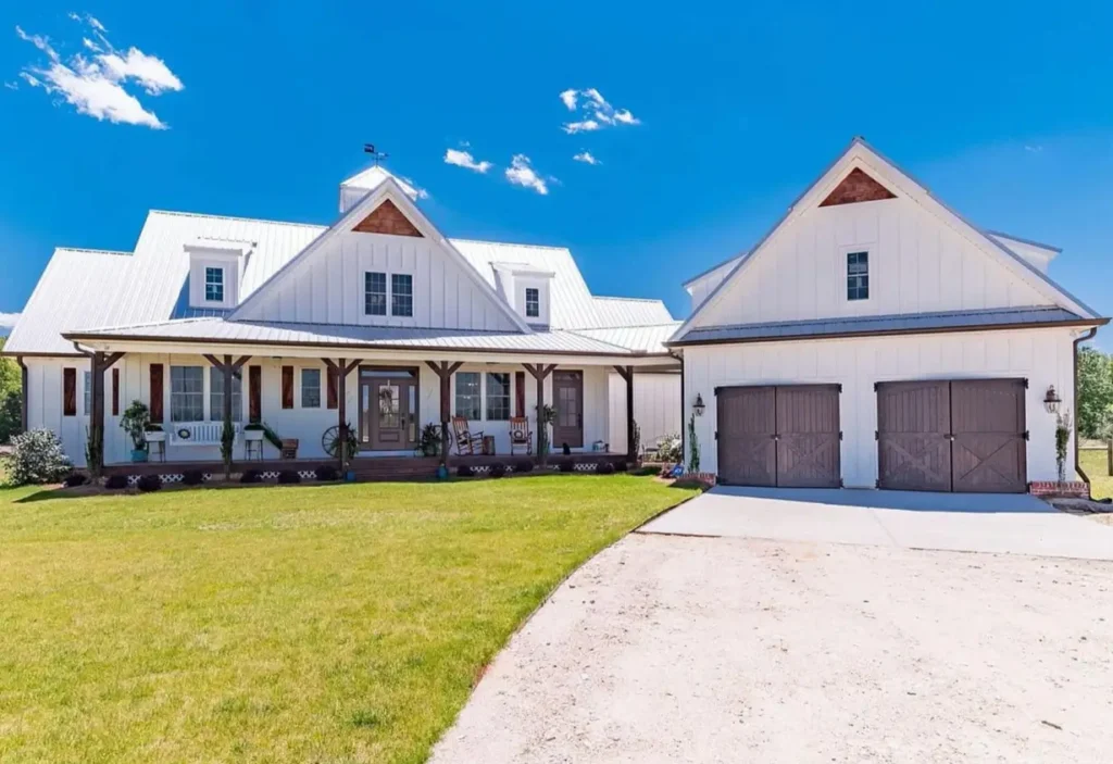 A sprawling modern white farmhouse exterior with dark wood shutters, porch columns, and matching carriage-style garage doors under a bright white metal roof.