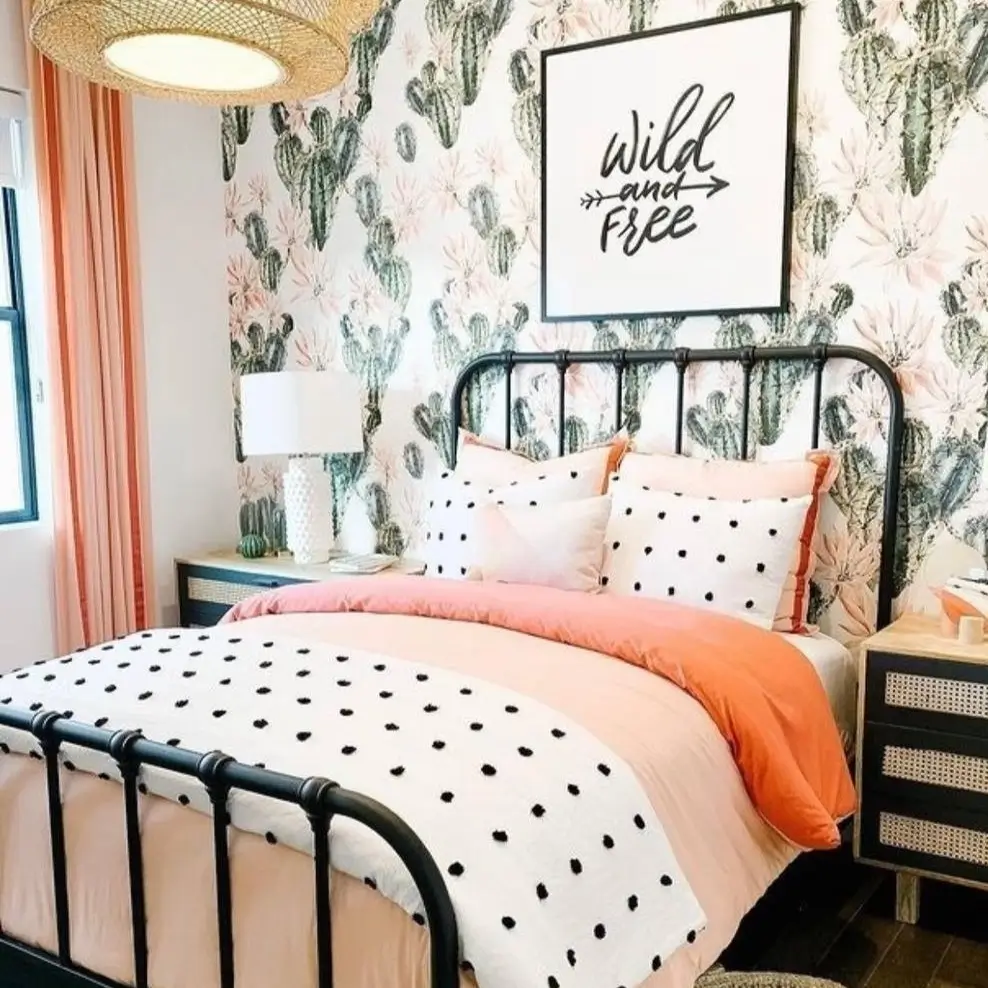 A bedroom with cactus-print wallpaper and a framed "Wild and Free" art print hanging above a black metal bed with pink and white bedding.