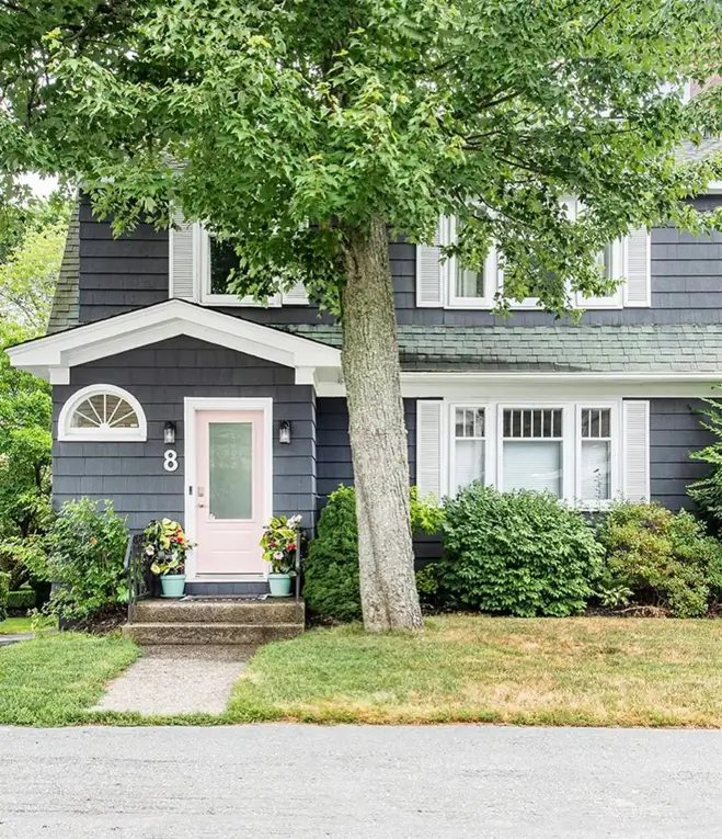 A dark gray shingle house with white shutters and a light pink front door.